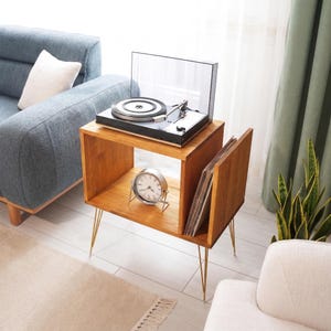 Op de afbeelding: Een houten platenspelerstandaard met een platenspeler, klok en vinylplaten. De standaard heeft een natuurlijke houten afwerking, gouden poten en een compartiment voor het opbergen van platen. Een blauwe bank en een wit kussen staan op de achtergrond.