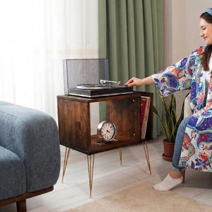 May include: A dark brown wooden record player stand with gold-colored hairpin legs. The stand has an open shelf with a silver clock and records. A black record player sits on top.