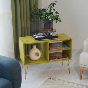 Puede incluir: Un mueble tocadiscos de madera verde lima con patas doradas de horquilla. El mueble tiene dos estantes, uno con discos de vinilo y otro con un jarr&oacute;n decorativo. Un tocadiscos negro se encuentra en la parte superior, con una planta en maceta en el fondo.