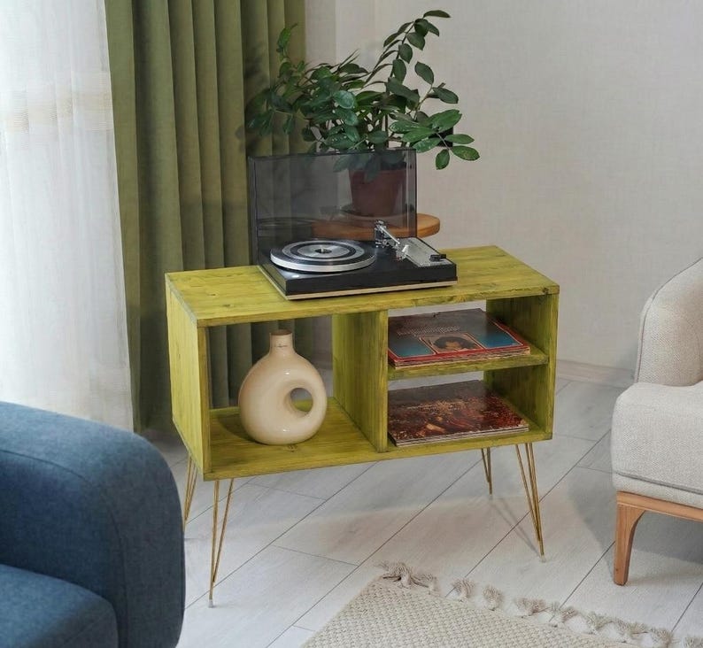 Puede incluir: Un mueble tocadiscos de madera verde lima con patas doradas de horquilla. El mueble tiene dos estantes, uno con discos de vinilo y otro con un jarr&oacute;n decorativo. Un tocadiscos negro se encuentra en la parte superior, con una planta en maceta en el fondo.