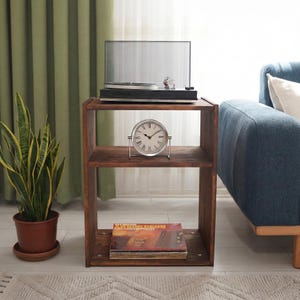 Puede incluir: Una mesa auxiliar de madera marrón oscuro con un tocadiscos encima, un reloj plateado en el estante del medio y discos de vinilo en el estante inferior. Una planta en maceta está a la izquierda. La mesa mide 61 cm de alto.