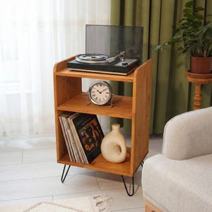 May include: A wooden record player stand with a black turntable on top, a silver clock, and a stack of vinyl records. The stand has three shelves and black hairpin legs. A white vase sits on the bottom shelf. The stand is made of light brown wood.