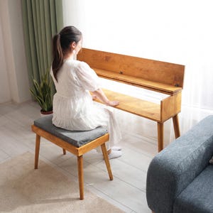 Könnte beinhalten: Eine Holz-Klavierbank mit einem grauen Kissen und einem passenden Holzklavier. Das Klavier hat eine aufklappbare Oberseite. Bank und Klavier sind aus hellem Holz gefertigt. Eine Person sitzt auf der Bank.