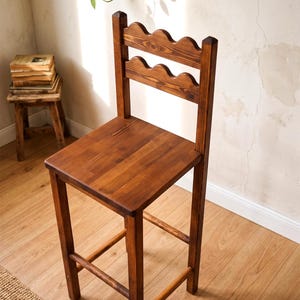 May include: A tall wooden bar stool with a dark brown finish, featuring a square seat and a backrest composed of two horizontal slats with a scalloped, wavy top edge. The stool has four straight legs connected by horizontal support stretchers.