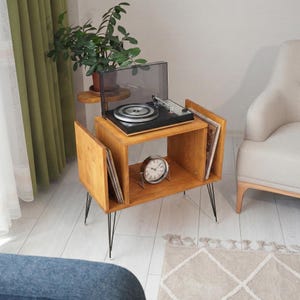 Puede incluir: Un mueble tocadiscos de madera con un tocadiscos, almacenamiento para discos de vinilo y un reloj. El mueble es de madera clara con patas de metal negro. Una planta en maceta se encuentra en un estante pequeño.
