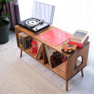 May include: A wooden media console with four tapered legs and two open storage compartments. The top surface holds a black record player, a pink vinyl record sleeve titled Dancing, and a stack of books. A red telephone booth model sits inside.
