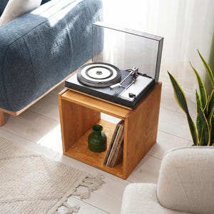 Può includere: Un giradischi nero con una copertura trasparente è posto su un tavolino a cubo in legno. Il giradischi ha un braccio argentato e un disco bianco e nero. Un vaso verde e dischi in vinile sono visibili sul ripiano del tavolo. La stanza ha un'estetica moderna.