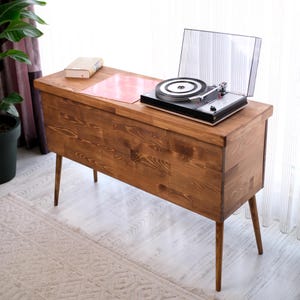 May include: A rectangular wooden storage console with a medium brown finish and four tapered legs. A black turntable with a clear plastic dust cover sits on the top surface, alongside a closed book and a square pink record sleeve.