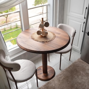 Può includere: Un tavolo da pranzo rotondo in legno con finitura marrone scuro, sostenuto da una base a piedistallo. Due sedie imbottite beige sono posizionate attorno al tavolo. Un albero di tazze in legno contiene diverse tazze beige e un sottobicchiere intrecciato è sul tavolo.