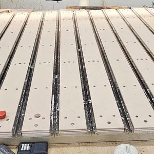 Puede incluir: Una base de máquina CNC que presenta una serie de listones de madera de color claro paralelos con orificios pretaladrados. Varios pomos circulares de color rojo brillante están insertados en las ranuras del lado izquierdo. Incluye rieles laterales negros.