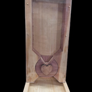 Könnte beinhalten: Ein hölzernes Vogelhaus mit einer transparenten Frontplatte und einem herzförmigen Akzent. Der Futterautomat ist aus hellfarbenem Holz gefertigt und hat eine kleine Schale am Boden zur Saatgutsammlung. Das Design ist schlicht und rustikal, für den Außenbereich geeignet.