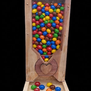 May include: A wooden candy dispenser with a clear front panel filled with multi-colored M&M's candies. The dispenser features a heart-shaped wooden knob in the center and a small tray at the base containing several loose M&M's candies.
