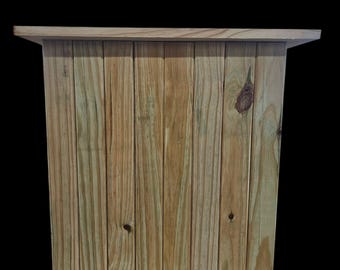 May include: A wooden planter box with a natural finish. The box is constructed from vertical wooden planks and features a simple, rectangular design. The planter has a flat top and sits on small wooden feet.