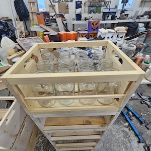 Puede incluir: Una caja de madera con varios frascos de vidrio en su interior. La caja está hecha de madera sin terminar y tiene un aspecto rústico. Los frascos son de vidrio transparente y están dispuestos en filas. La caja está sentada en un estante de madera.