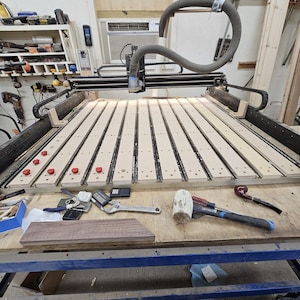 May include: A close-up of a CNC machine with a wooden work surface. The machine has a black and gray body with a gray hose attached to it. The work surface is made of plywood and has a series of parallel grooves running across it. There are several red buttons on the surface, as well as a few tools, including a hammer, a wrench, and a pipe.