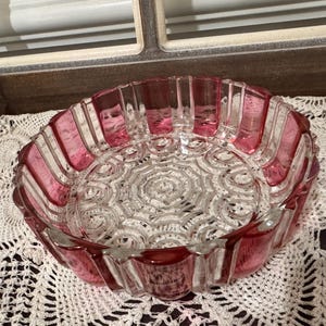 May include: A clear glass bowl with a pink rim and vertical pink stripes. The bowl has a decorative, circular pattern in the center. The bowl is sitting on a white, crocheted doily.