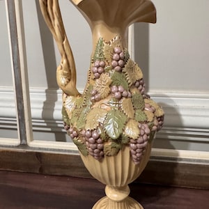 May include: A beige ceramic pitcher with a decorative handle and a wide, scalloped rim. The pitcher is adorned with sculpted green leaves and clusters of purple grapes. The base is a pedestal design.