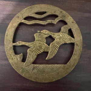 May include: A round, antique brass trivet featuring a detailed relief of two flying ducks against a stylized sky. The trivet has a circular frame with cut-out details and a weathered, aged appearance, suggesting vintage craftsmanship. The ducks are in mid-flight.