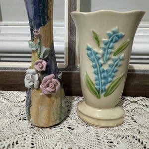 May include: Two ceramic vases. One is tall and slender, with a textured surface and applied pink and white roses. The other is cream-colored, with blue floral designs and green leaves. Both vases are on a white lace surface.