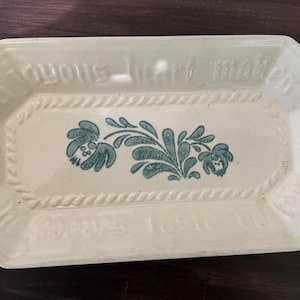 May include: Cream-colored rectangular ceramic serving dish with an octagonal shape. The dish features a teal floral design in the center and embossed text around the edges. A rope-like border frames the central design.