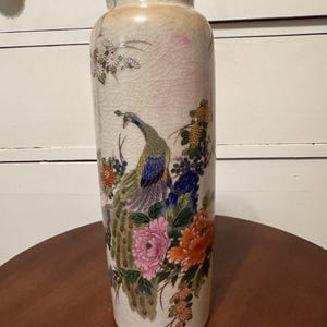 May include: A tall, off-white ceramic vase with a crackle glaze finish. The vase features a painted peacock with colorful plumage, surrounded by pink, orange, and blue flowers. The vase is a decorative item.