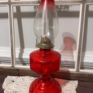May include: A vintage red glass oil lamp with a clear glass chimney. The lamp has a textured base and a brass burner. The top of the lamp is a deep red that fades to clear. The lamp is sitting on a white lace doily.