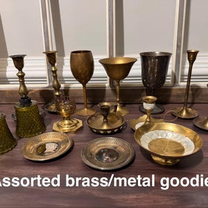 May include: An assortment of brass and metal decorative items, including candlesticks, goblets, and small dishes. The items are in various shapes and sizes, with a warm, antique aesthetic. The text "Assorted brass/metal goodies" is at the bottom.