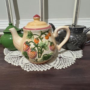 May include: Three teapots on a lace doily. The central teapot is cream-colored with a strawberry design. A green teapot is to the left, and a silver teapot is to the right. The teapots are on a dark wooden surface.