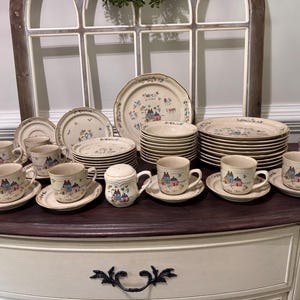 May include: A large ceramic dinnerware set featuring a cream-colored base with a repeating pattern of small houses, trees, and floral accents in blue, pink, and green. The collection includes stacked dinner plates, salad plates, bowls, and mugs.