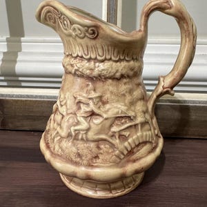 May include: Tan ceramic pitcher with a detailed relief design. The pitcher features a curved spout, a large handle, and a decorative base. The relief depicts a hunting scene with figures on horseback and a bridge. A vintage-style decorative piece.
