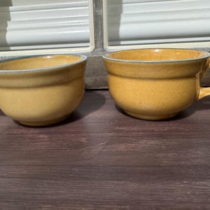 May include: Two yellow ceramic mugs with handles. The mugs have a rounded shape and a slightly textured surface. The rims are accented with a thin, light blue line. The mugs are placed on a dark wooden surface.