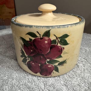 May include: A ceramic apple-themed cookie jar with a lid. The jar is decorated with a cluster of red apples with green leaves on a cream background. The lid is a light cream color with a knob.
