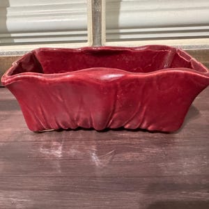 May include: A rectangular, red ceramic planter with a ruffled edge. The planter has a glossy finish and is designed for holding plants. The container is sitting on a wooden surface.