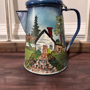 May include: A vintage blue enamelware coffee pot with a hand-painted landscape scene. The scene depicts a white cottage with a red chimney, a stone path, and lush greenery. The pot has a blue lid and handle.