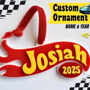 May include: A custom ornament with a red and yellow flame design. The name "Josiah" is in yellow, with the year "2025" in a red oval. A red ribbon is attached for hanging. The text "Custom Ornament NAME & YEAR" is also visible.