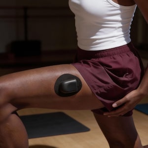 Puede incluir: Un dispositivo médico negro está adherido al muslo con un parche adhesivo circular. La persona lleva pantalones cortos granates y una camiseta sin mangas blanca. El fondo está borroso, lo que sugiere un entorno interior.