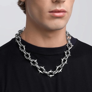 Puede incluir: Un collar de cadena plateada con un diseño puntiagudo. El collar está hecho de eslabones gruesos e interconectados y presenta púas afiladas y puntiagudas a lo largo de los bordes exteriores. El collar se usa alrededor del cuello y descansa sobre una camiseta negra.