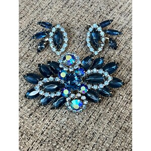 May include: A vintage blue and clear rhinestone brooch and earrings set. The brooch features a cluster of blue and clear rhinestones in a floral design. The earrings are simple and feature a single blue rhinestone.