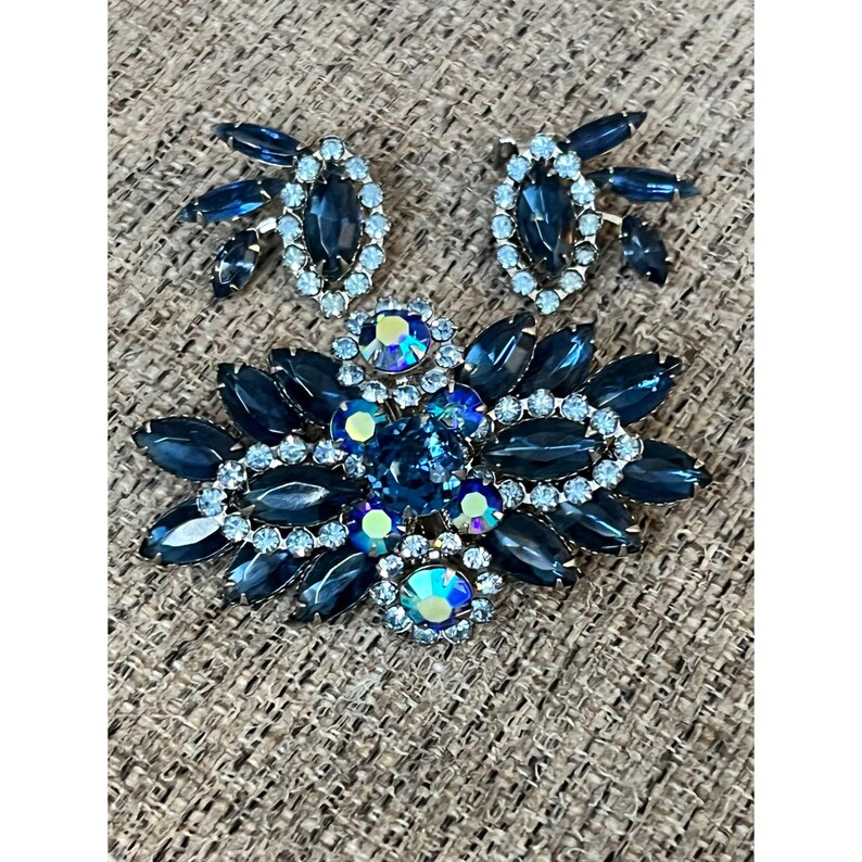 May include: A vintage blue and clear rhinestone brooch and earrings set. The brooch features a cluster of blue and clear rhinestones in a floral design. The earrings are simple and feature a single blue rhinestone.