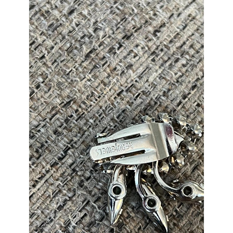 May include: A silver tone brooch with a cluster of clear and green rhinestones. The back of the brooch has a metal clasp and the words "Benrus Jewels" engraved on it.