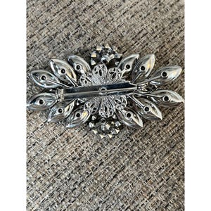 May include: The back of a silver brooch with a floral design. The brooch is decorated with clear rhinestones and has a pin and clasp closure.