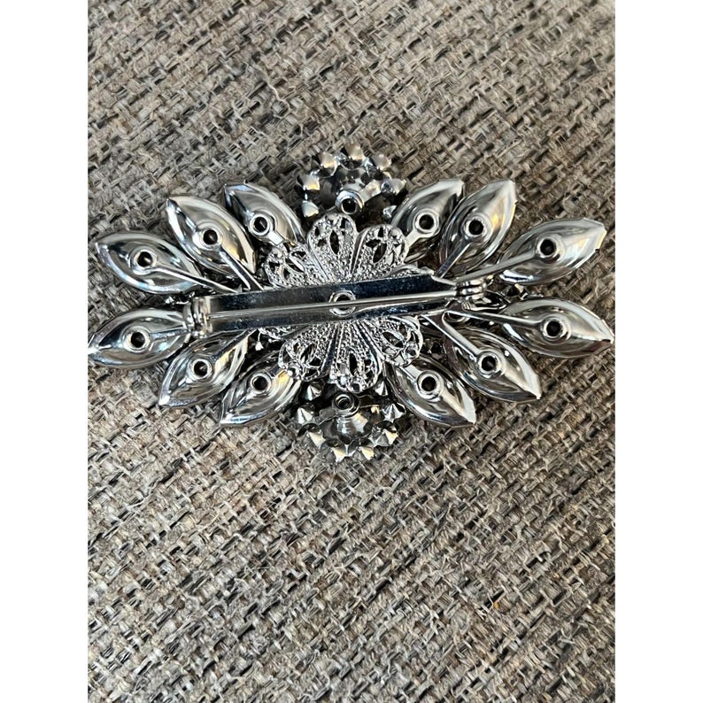 May include: The back of a silver brooch with a floral design. The brooch is decorated with clear rhinestones and has a pin and clasp closure.