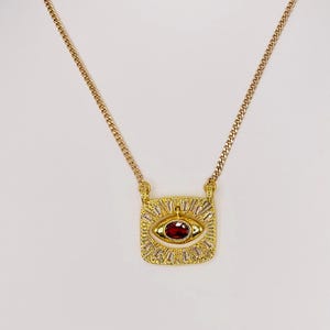 May include: Gold-coloured necklace with a rectangular pendant featuring an eye design. The eye has a red centre stone and is surrounded by clear, baguette-cut stones. The chain is a delicate curb link style.