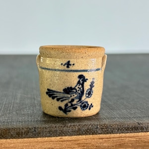 May include: Small, tan stoneware crock with a blue bird and floral design. The miniature crock has a cylindrical shape with two small handles and a light brown rim. A blue line and dots encircle the crock near the rim.
