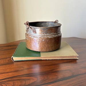 May include: A small, antique copper bucket with a handle, resting on a green and tan book. The bucket has a weathered appearance, with visible signs of age and use. The background is a wooden table and a neutral wall.