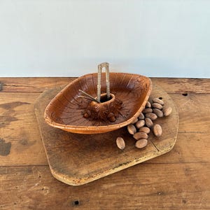 Puede incluir: Un juego de cascanueces de madera vintage con un cuenco marrón tallado con hojas y un cascanueces de metal. El juego está sobre una tabla de cortar de madera, con varias nueces esparcidas. Un tema rústico y otoñal.