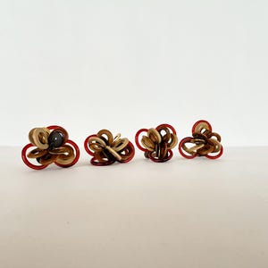 May include: Four abstract flower-shaped earrings. Each earring features a combination of red, tan, and brown circular elements. One earring has a dark gray center. The earrings are arranged in a row on a white surface.