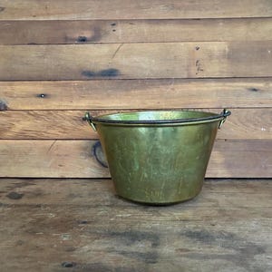 May include: A vintage brass bucket with a slightly tapered shape and two small handles. The bucket has a tarnished, aged appearance, suggesting it is an antique. It sits on a wooden surface with a wooden plank background.