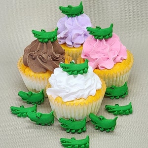 Peut inclure: Quatre cupcakes décorés de dessus d'alligator verts. Les cupcakes sont glacés en rose, violet, blanc et chocolat. Les cupcakes sont sur une surface beige.