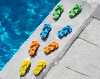 Può includere: Una collezione di infradito in miniatura colorate in verde, blu, arancione e giallo con motivi a pois. I sandali sono disposti su una superficie grigio chiaro accanto a una piscina di acqua blu, evocando un tema estivo.